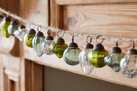 The Ekiti Bauble Garland - Mixed Green & Clear by nkuku features small recycled glass ornaments in green, clear, and silver, with round and ribbed shapes, all hanging from twine—ideal for rustic festive décor against a light wood background.