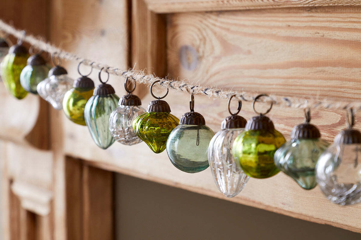 The Ekiti Bauble Garland - Mixed Green & Clear by nkuku features small recycled glass ornaments in green, clear, and silver, with round and ribbed shapes, all hanging from twine—ideal for rustic festive décor against a light wood background.