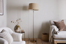 A cozy living room with a neutral palette features the nkuku Ehadi Antique Floor Lamp in antique brass, a white armchair, side table with dried flowers in a glass vase, beige sofa with pillows and throw, and a woven basket on the wooden floor.