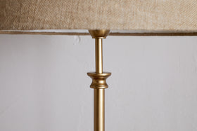 A close-up of the nkuku Ehadi Antique Floor Lamp in antique brass, featuring a slender metallic top and decorative collar with a textured beige shade, set against a plain white wall for a minimalist look.