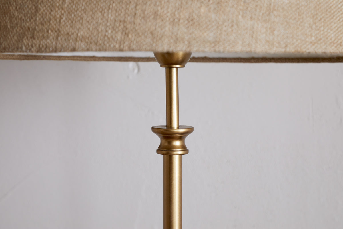 A close-up of the nkuku Ehadi Antique Floor Lamp in antique brass, featuring a slender metallic top and decorative collar with a textured beige shade, set against a plain white wall for a minimalist look.