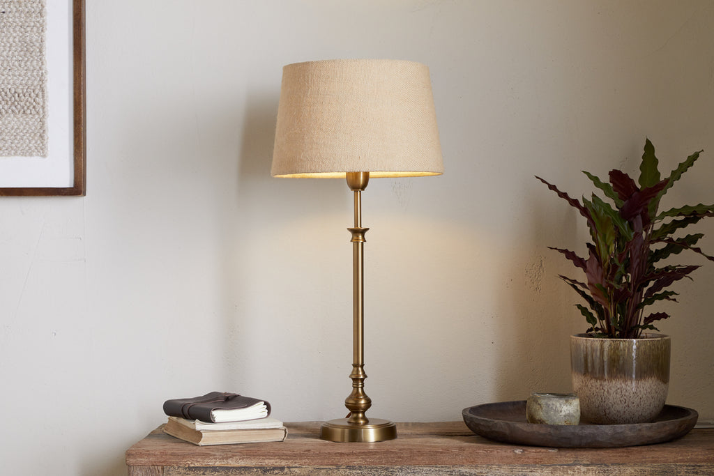 The nkuku Ehadi Antique Table Lamp - Antique Brass, with a beige fabric shade, sits on a wooden table beside books, a plant, a candle, and a tray against a light-colored wall.