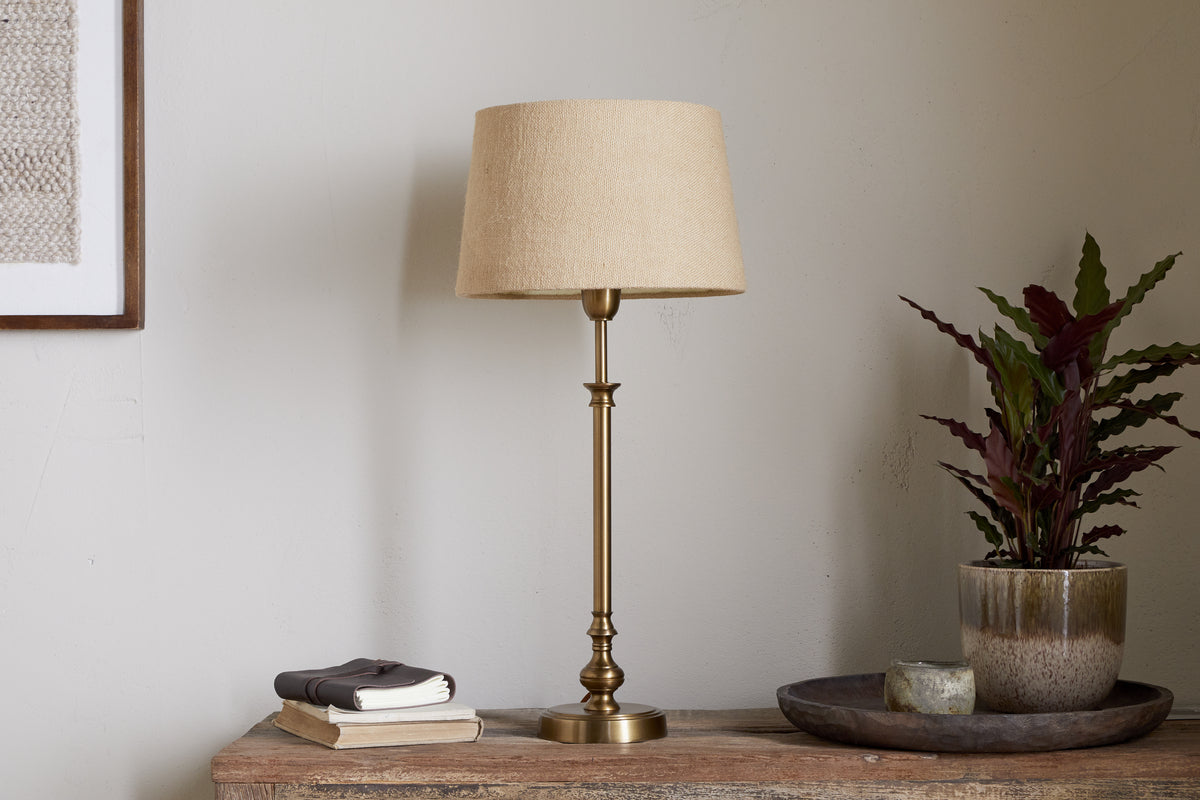 The nkuku Ehadi Antique Table Lamp in antique brass sits elegantly on the table, bringing a stylish and classic touch to your space.