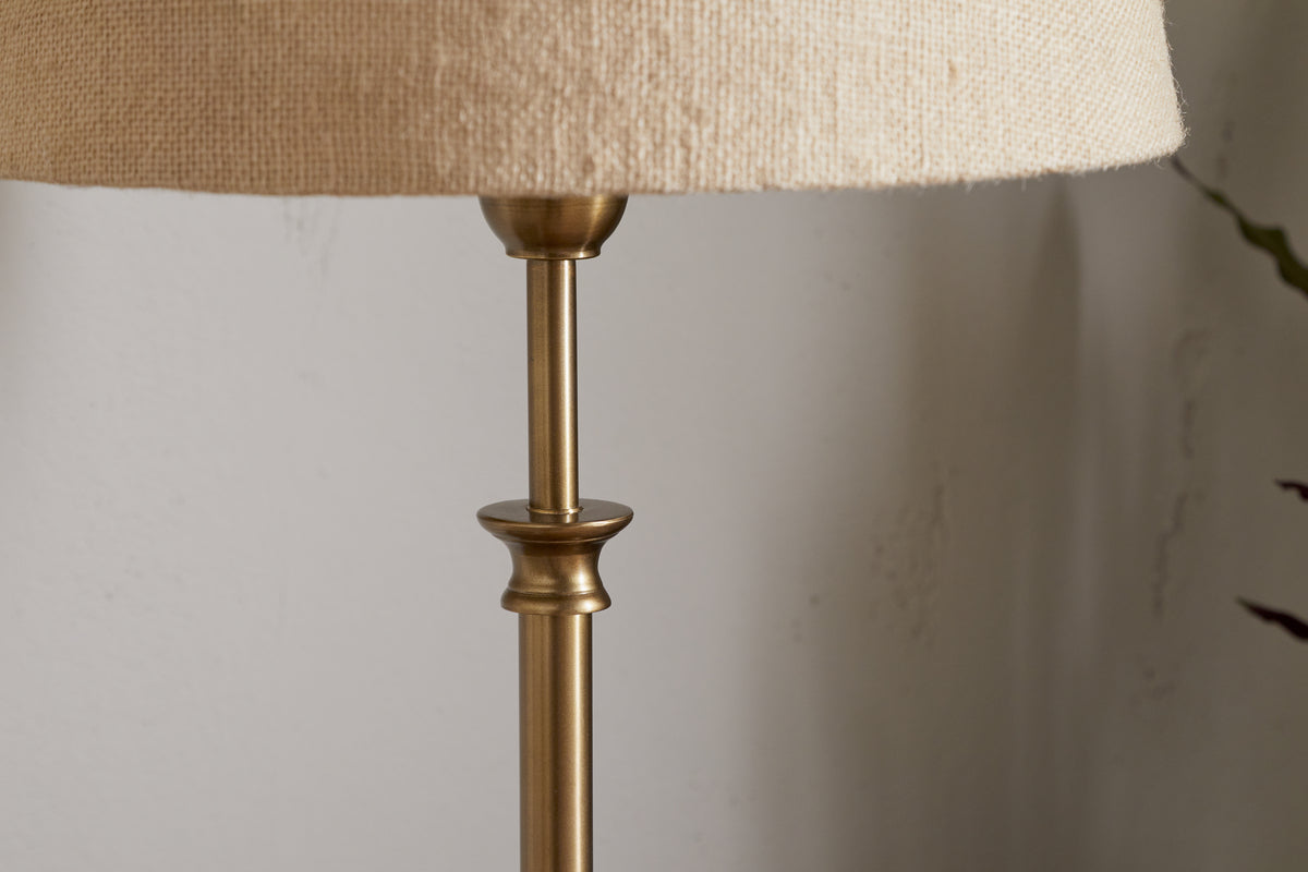 A close-up shows the upper part of the nkuku Ehadi Antique Table Lamp in antique brass, featuring a beige textured fabric shade against a light-colored wall.