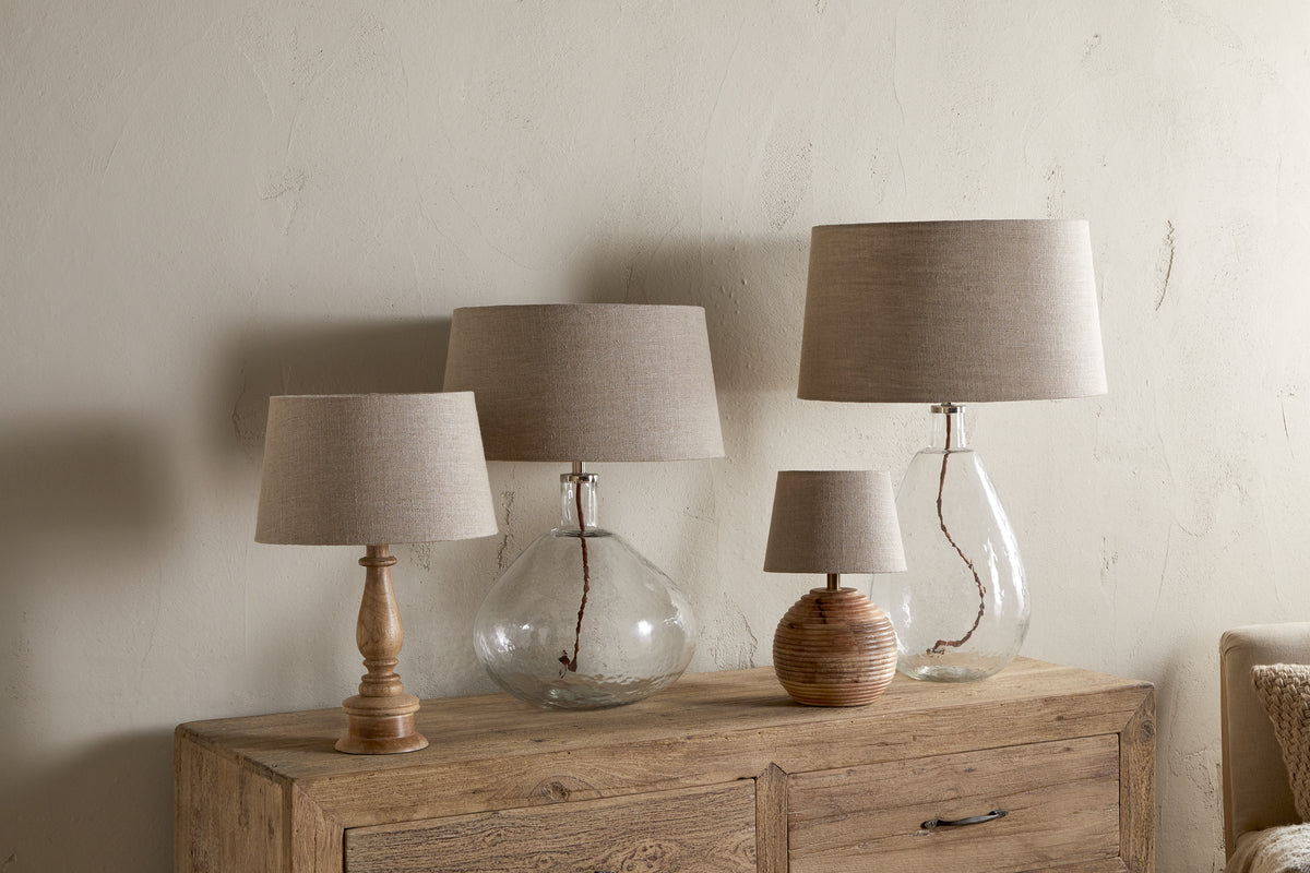 Four Embaata Linen Lampshades - Natural by nkuku, with textured linen, sit on a wooden sideboard—two have clear glass bases, one a wooden base, and another a ribbed round base. The neutral wall highlights their artisanal look.