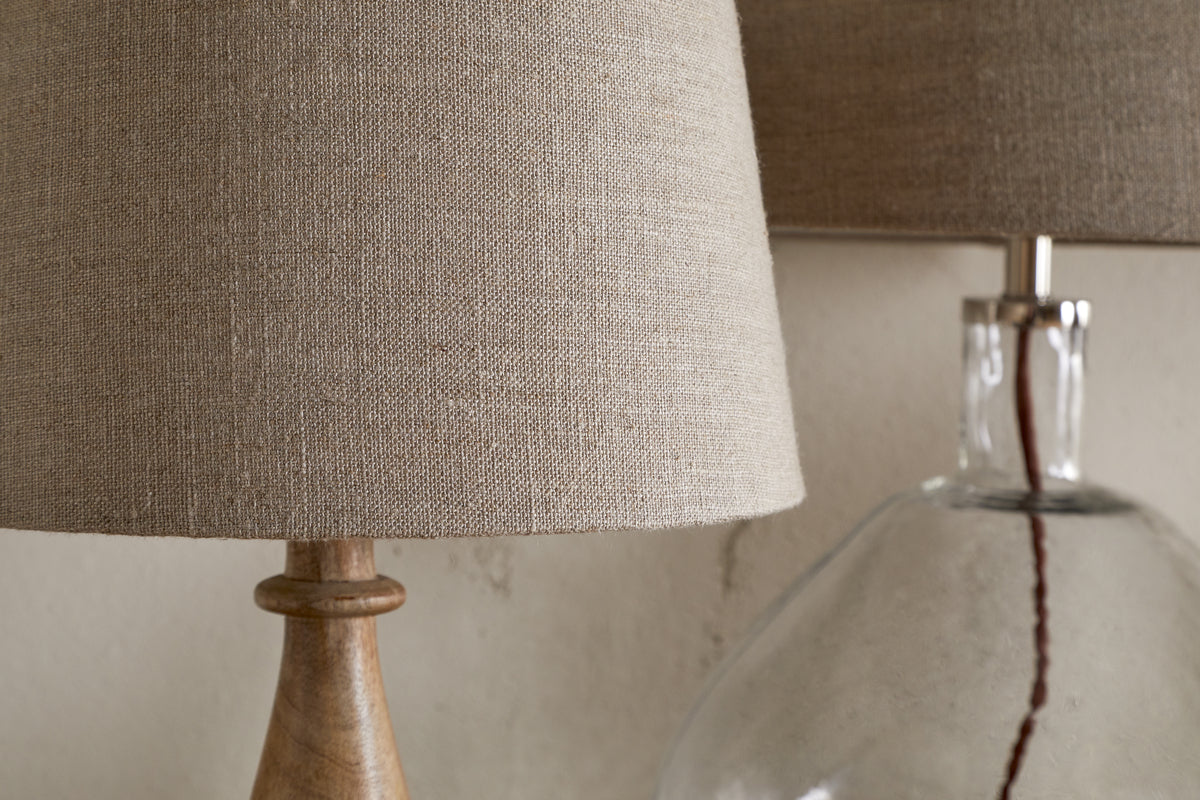 Close-up of two nkuku Embaata Linen Lampshade - Natural lamps: one with a wooden base, the other with a clear glass base—both perfect as bedroom lights against a neutral wall.
