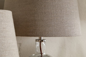 A close-up of the nkuku Embaata Linen Lampshade - Natural, featuring a textured linen shade and transparent glass base, sits beside a similar bedroom lamp against a neutral wall.