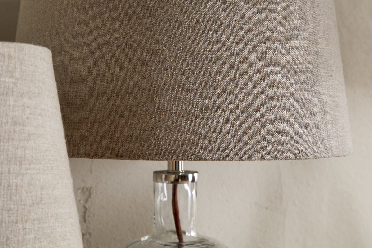 A close-up of the nkuku Embaata Linen Lampshade - Natural, featuring a textured linen shade and transparent glass base, sits beside a similar bedroom lamp against a neutral wall.