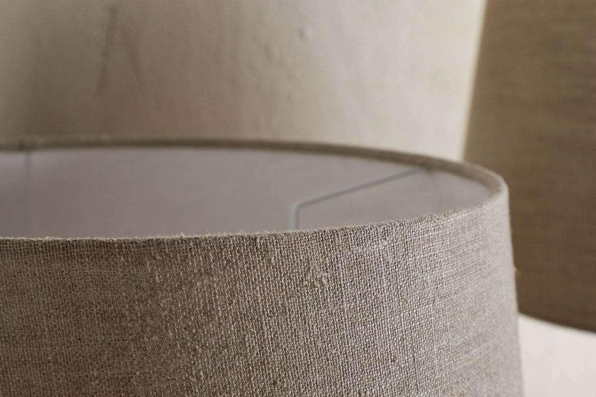 Close-up of the nkuku Embaata Linen Lampshade in Natural, highlighting its beige textured linen and inner metal frame. A second lampshade is blurred in the background, ideal for soft bedroom lighting against a neutral wall.