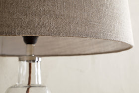 Close-up of the nkuku Embaata Linen Lampshade - Natural, featuring a textured linen shade and clear glass base, ideal for bedroom lighting, set against a light, neutral background.