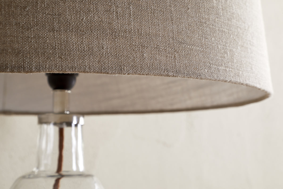 Close-up of the nkuku Embaata Linen Lampshade - Natural, featuring a textured linen shade and clear glass base, ideal for bedroom lighting, set against a light, neutral background.
