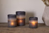 Three nkuku Etti Tealight Holders in navy and smoke cast a warm glow on a rustic wooden surface, accompanied by a beige textured vase with dried flowers, all set against a neutral wall backdrop.