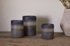 Three Etti Tealight Holders in navy and smoke by nkuku, each a different height, are displayed on a rustic wooden surface beside a textured vase with dried flowers, creating an ambient glow against a neutral wall.