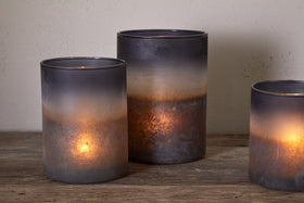 Three Etti Tealight Holders - Navy & Smoke by nkuku, each with lit candles, sit on a rustic wooden surface. Their smoky ombre glass fades from dark gray rims to warm amber bases, casting an ambient glow.