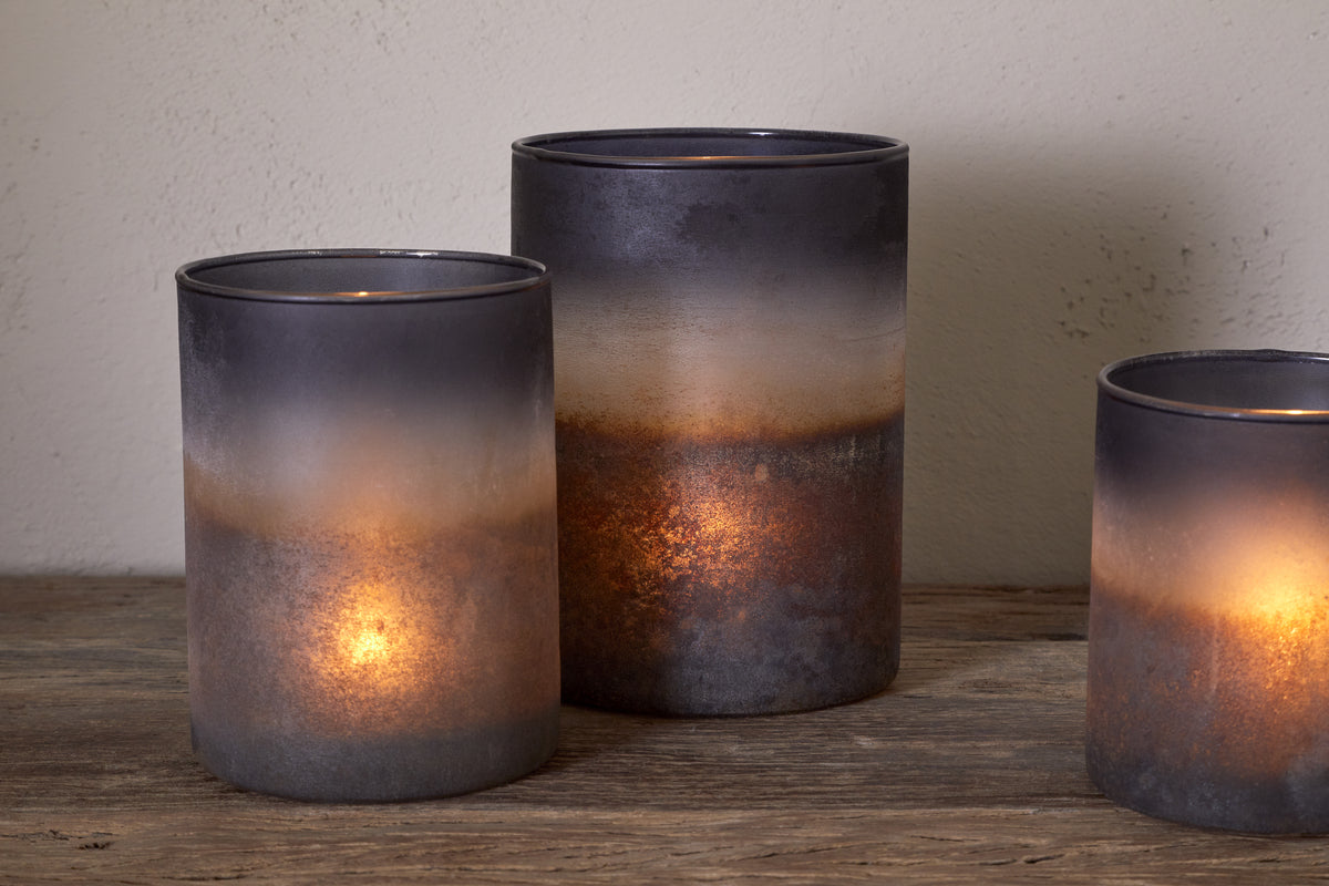 Three Etti Tealight Holders - Navy & Smoke by nkuku, each with lit candles, sit on a rustic wooden surface. Their smoky ombre glass fades from dark gray rims to warm amber bases, casting an ambient glow.