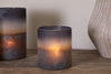 The nkuku Etti Tealight Holders in Navy & Smoke, one lit and one unlit, sit on a wooden surface beside a textured vase, casting an inviting ambient glow.