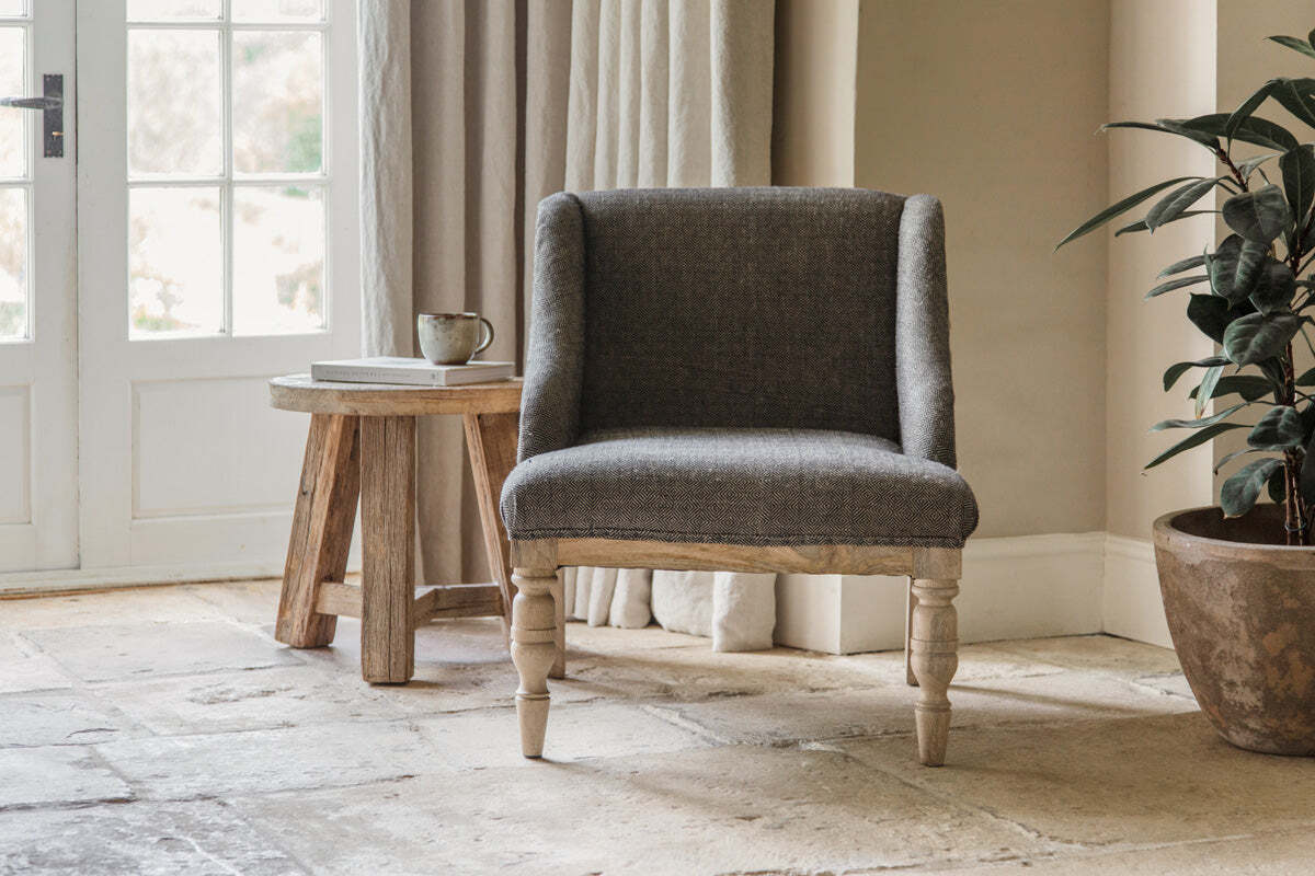 The nkuku Elbu Deconstructed Jacquard Weave Armchair in Charcoal with wooden legs sits on a stone tile floor next to a rustic mango wood side table holding a mug and book, while sunlight streams through glass-paneled double doors.