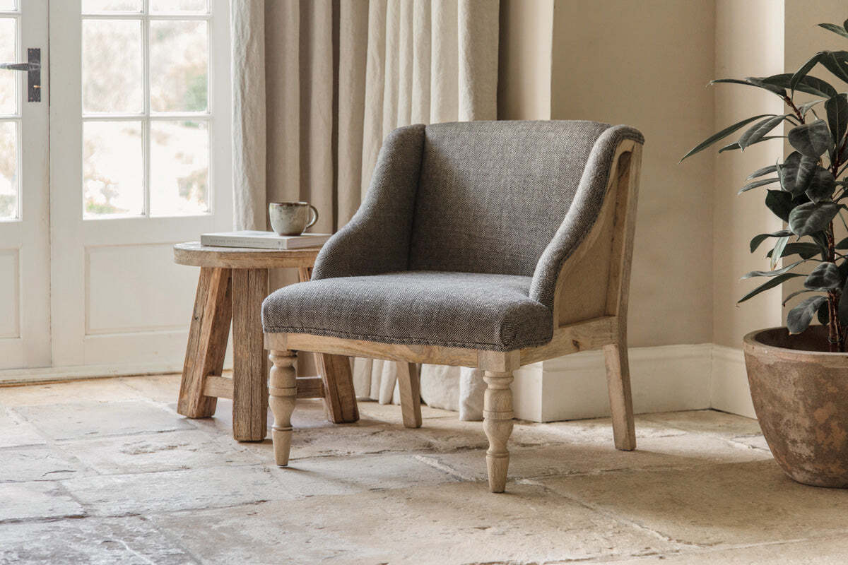 A cozy nkuku Elbu Deconstructed Jacquard Weave Armchair in Charcoal with wooden legs rests on stone tiles beside a rustic mango wood side table holding a mug and book. Sunlight pours through French doors, highlighting the neutral-toned space and potted plant.