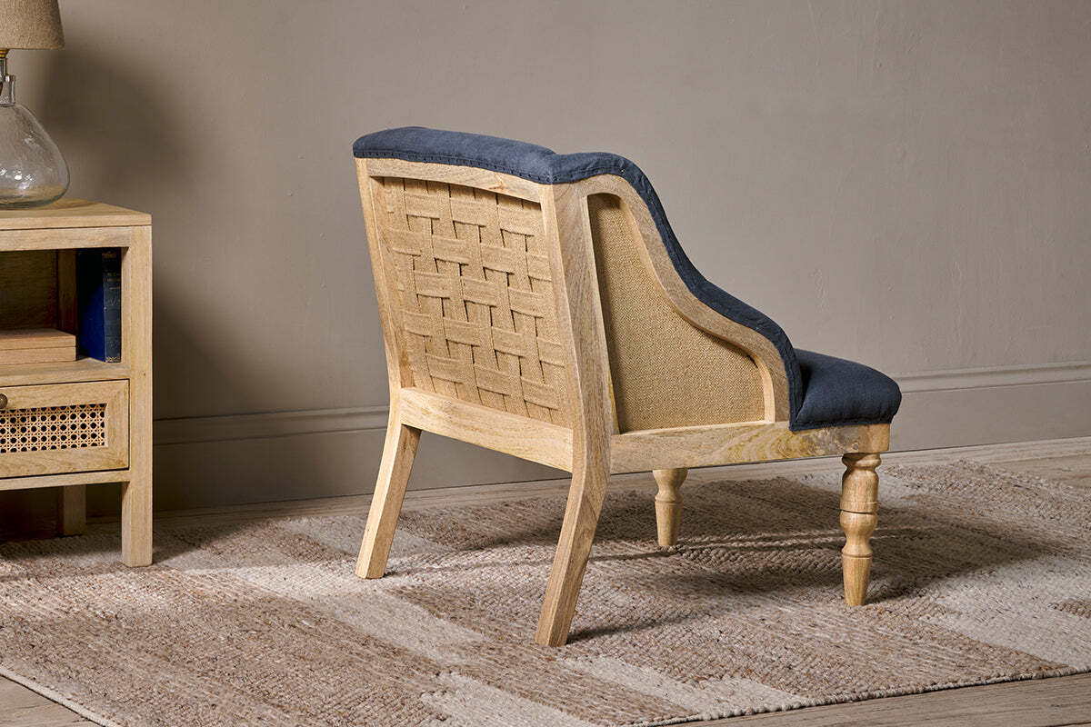 The Elbu Deconstructed Linen Armchair by nkuku, featuring a woven cane back and charcoal linen seat, sits on a beige rug. To the left, a mango wood side table holds a glass vase and books against a light gray wall.