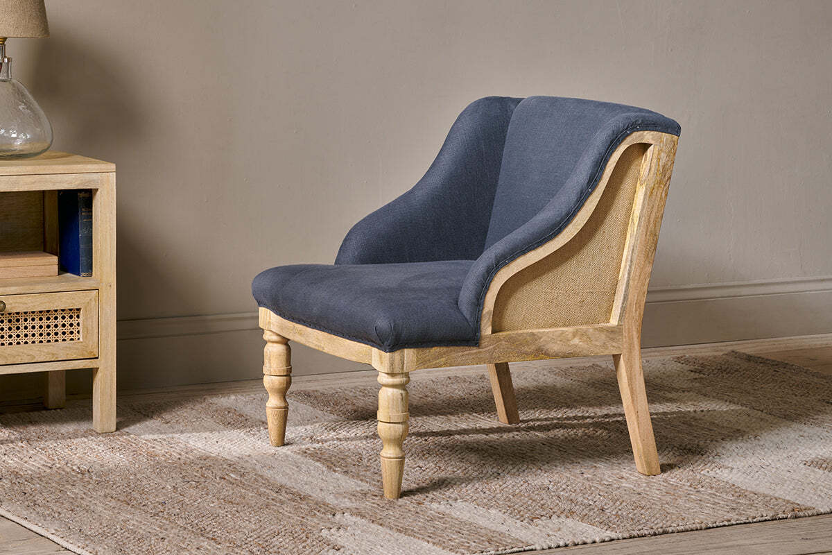 The nkuku Elbu Deconstructed Linen Armchair in charcoal sits on a beige patterned rug beside a light mango wood nightstand topped with books and a glass lamp, set against a neutral-colored wall.