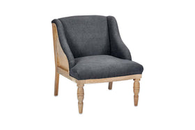 The Elbu Deconstructed Linen Armchair by nkuku features a curved backrest, armrests, and dark charcoal linen upholstery. Its light mango wood legs have carved detailing in front and simple back legs. Shown on a white background.