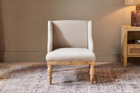 The nkuku Elbu Deconstructed Linen Armchair in Stone features a curved back and nailhead trim, wooden legs, and sits on a light patterned rug. Behind it: a tan wall, natural mango wood side table, and brown ceramic base lamp.