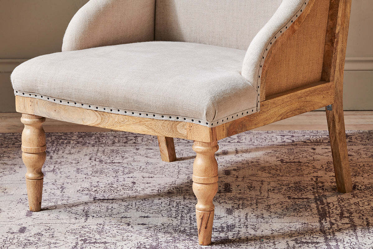 The Elbu Deconstructed Linen Armchair - Stone by nkuku features beige linen upholstery, a natural mango wood frame and legs, silver nailhead trim, and gently curved armrests. Shown on a muted patterned rug against a light wall.