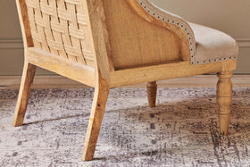 The Elbu Deconstructed Linen Armchair - Stone by nkuku features a natural mango wood frame, woven backrest, light beige linen upholstery, decorative nailhead trim, and turned front legs—perfect for a warm, neutral space.