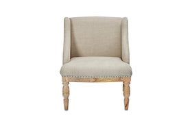 The nkuku Elbu Deconstructed Linen Armchair - Stone features beige linen upholstery, a curved backrest with winged sides, light mango wood turned legs, and nailhead trim along the bottom edge. It is shown against a plain white background.