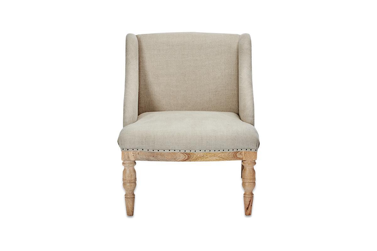 The nkuku Elbu Deconstructed Linen Armchair - Stone features beige linen upholstery, a curved backrest with winged sides, light mango wood turned legs, and nailhead trim along the bottom edge. It is shown against a plain white background.