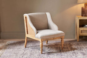 The Elbu Deconstructed Linen Armchair - Stone by nkuku, with light beige linen upholstery and a natural mango wood frame featuring turned front legs, sits on a patterned rug in a cozy room with neutral walls and wooden furniture.