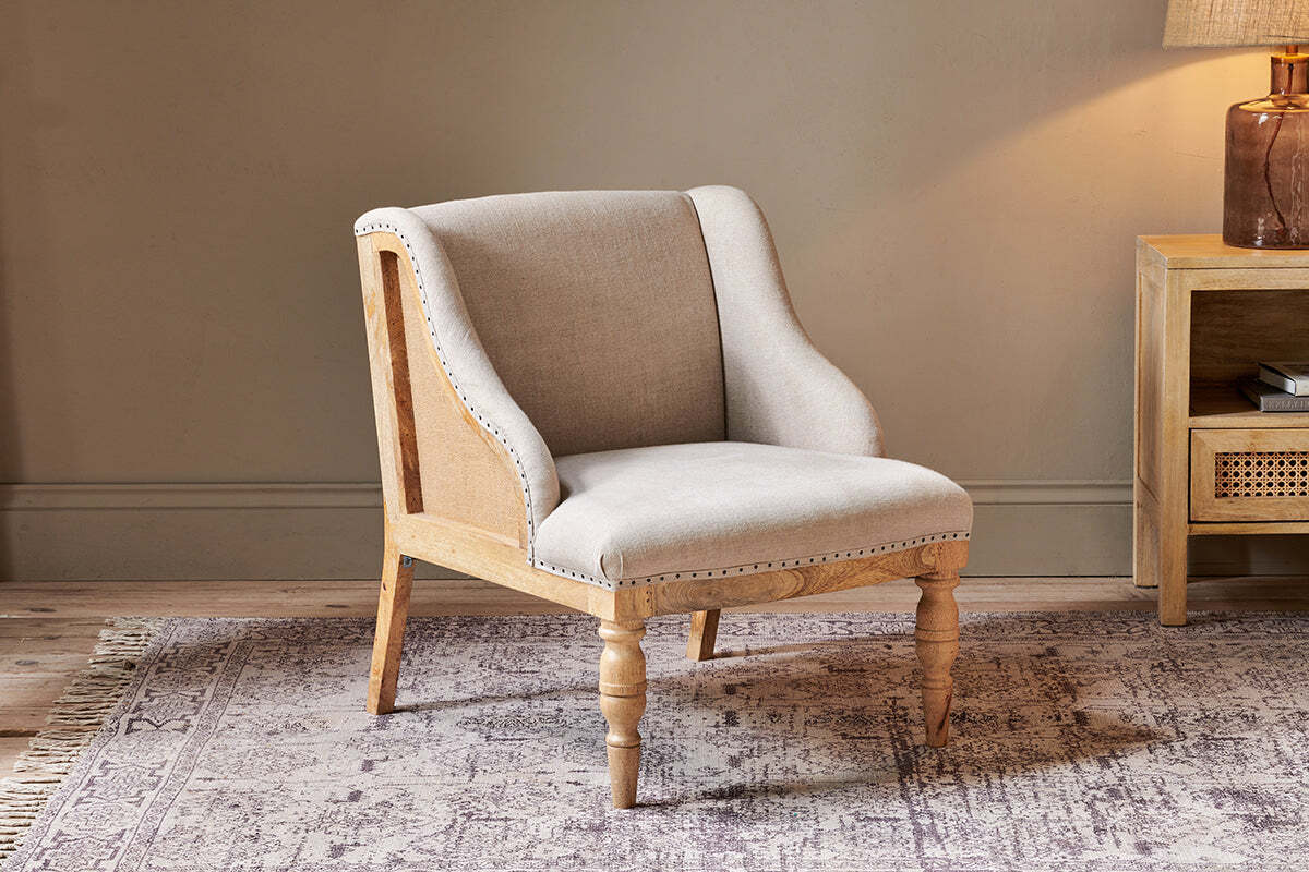The Elbu Deconstructed Linen Armchair - Stone by nkuku, with light beige linen upholstery and a natural mango wood frame featuring turned front legs, sits on a patterned rug in a cozy room with neutral walls and wooden furniture.