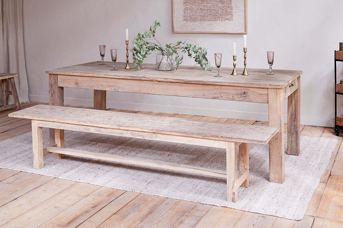 The Fioni Reclaimed Wood Bench by nkuku pairs with a rustic dining table atop a light woven rug. Candlesticks, goblets, and a glass vase with greenery complete the timeless, neutral farmhouse look.