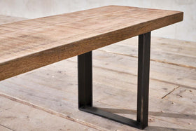 A close-up of nkuku's Fia Bench showcases its thick, rectangular mango wood seat with a natural finish and black metal angular legs, set on a rustic wooden floor against a light, textured wall.