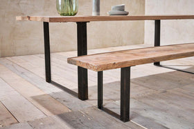 A wooden table and nkuku Fia Benches, with thick mango wood tops and black angular legs, stand on a weathered floor. Plates and glass vases rest atop the table, set against a beige textured wall in the background.