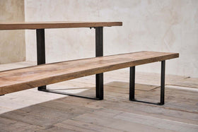 A close-up of the minimalist Fia Bench by nkuku, featuring a thick mango wood top and bold angular legs, set on rustic wooden floorboards against a textured light beige wall.