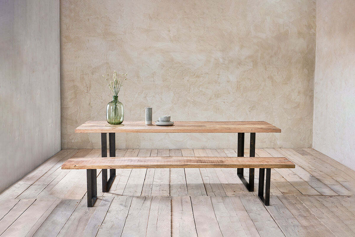 The nkuku Fia Bench, crafted from mango wood with angular legs, pairs with a matching table. On top: a green glass vase with dried flowers and ceramic dinnerware. The minimalist room features textured walls and wooden floors.
