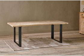 The nkuku Fia Dining Table, crafted from sustainable mango wood with a light natural finish and black iron base, stands on a textured beige rug over wooden floors against a light brown wall, with part of a wooden cabinet visible on the right.