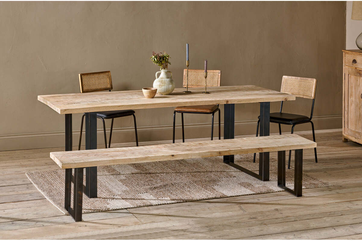 The Fia Dining Table by nkuku, crafted from sustainable mango wood with black metal legs, is styled with ceramics and flowers. Three wooden chairs and a matching bench surround the table on a textured rug in this neutral-toned space.