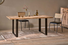 The Fia Dining Table by nkuku, crafted from sustainable mango wood with black metal legs, sits on a woven rug with cane-back chairs. Glass vases, dried flowers, and a small bowl adorn the table; a wooden cabinet stands against the beige wall.