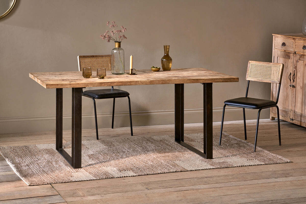 The Fia Dining Table by nkuku, crafted from sustainable mango wood with black metal legs, sits on a woven rug with cane-back chairs. Glass vases, dried flowers, and a small bowl adorn the table; a wooden cabinet stands against the beige wall.