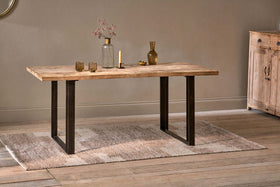 The nkuku Fia Dining Table, crafted from sustainable mango wood with black metal legs, stands on a textured beige rug and displays a glass vase of dried flowers, brass candle holder, gold bottle, and two low glasses on light wood floors.