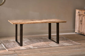 The Fia Dining Table by nkuku, featuring a rectangular mango wood top and black metal legs, stands on a beige woven rug in a room with light wood floors and neutral walls. A wooden cabinet is partially visible on the right.