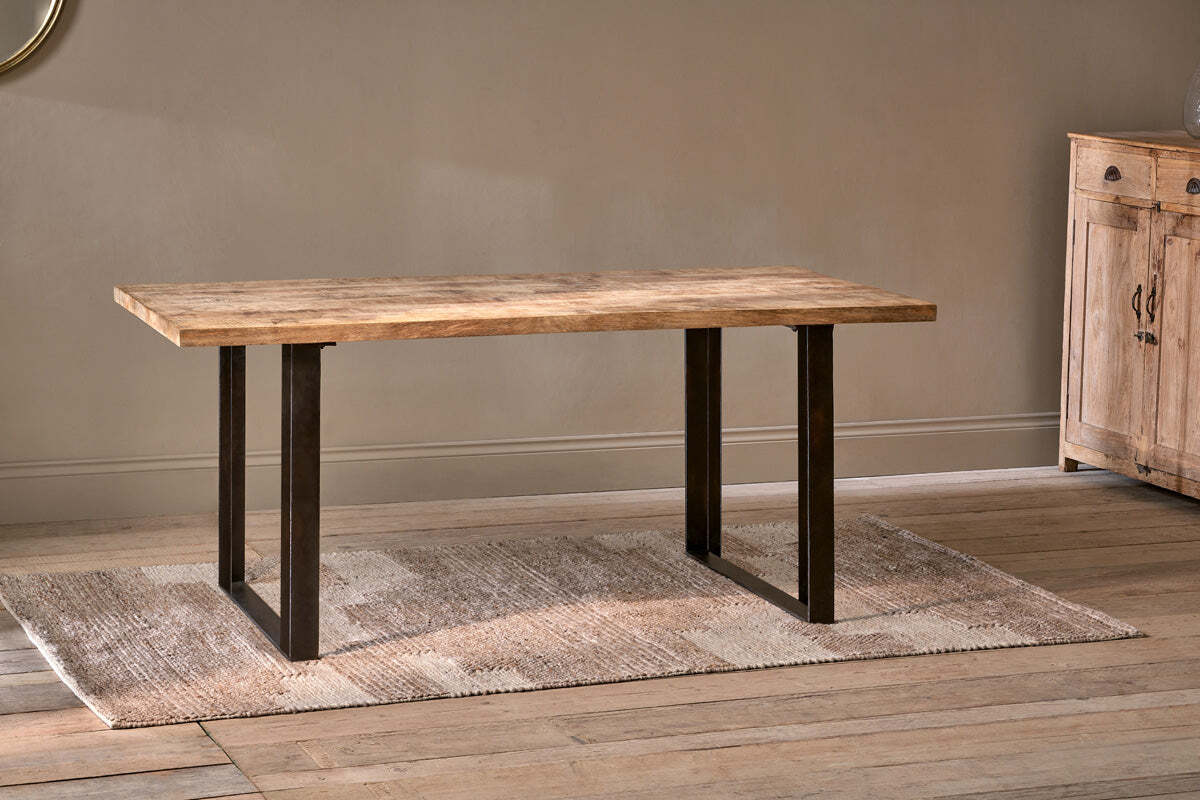 The Fia Dining Table by nkuku, featuring a rectangular mango wood top and black metal legs, stands on a beige woven rug in a room with light wood floors and neutral walls. A wooden cabinet is partially visible on the right.