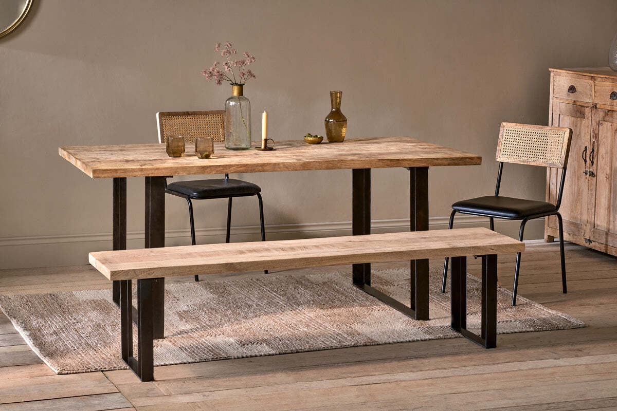 A dining space featuring the nkuku Fia Dining Table in sustainable mango wood with black metal legs, flanked by two cane-back chairs and a matching bench. The table is styled with glass vases, glasses, dried flowers; a sideboard and rug finish the look.