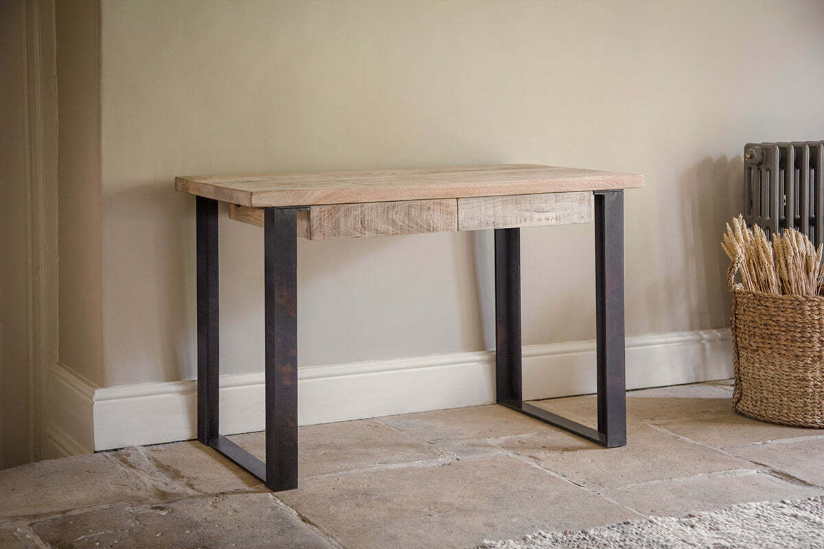 The nkuku Fia Mango Wood Desk - Natural, with metal legs and two drawers, stands against a wall on a stone floor. A basket of wheat sits to its right.