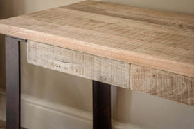 The nkuku Fia Mango Wood Desk - Natural features a slim mango wood tabletop with visible grain, paired with straight dark metal legs, shown against a beige wall and white baseboard.