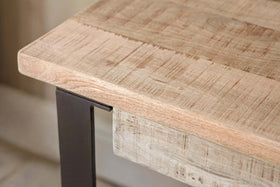 A close-up of the Fia Mango Wood Desk - Natural by nkuku shows its light mango wood with visible natural grain, supported by a slim black metal leg for a rustic, industrial style. The wood has an unfinished or lightly finished appearance.
