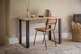 The nkuku Fia Mango Wood Desk - Natural features rustic charm, paired with a brown leather chair, a vase of dried flowers, books, and a basket of wheat stalks placed near a radiator.