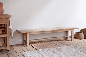 The nkuku Fioni Reclaimed Wood Bench in Natural, featuring a weathered finish, adds farmhouse charm on a plank floor against a white wall. Beside it sits a wooden cart with baskets.